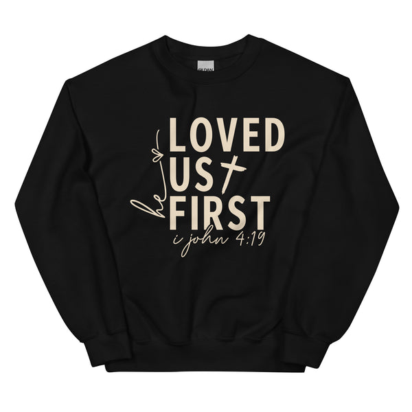 Black Christian sweatshirt with He Loved Us First 1 John 4:19 text on the front, laid flat on a neutral background.
